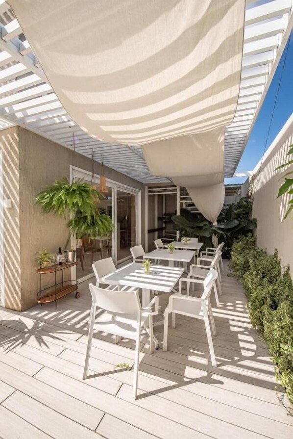 An outdoor patio with white tables and chairs, a large plant, and a pergola providing shade. - Paraíso Noronha (Fernando de Noronha)