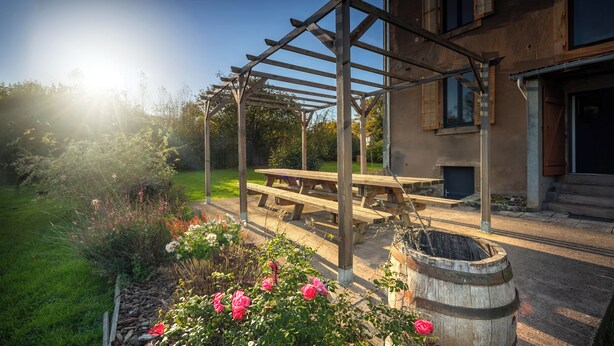 Une pergola en bois avec une table de pique-nique et un tonneau, située dans un coin du jardin où poussent des rosiers.
