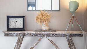 Une table en bois rustique, ornée d’une horloge et d’un vase de fleurs séchées, est placée devant un mur à la texture particulière, sur lequel est accroché un grand miroir en arc de cercle.