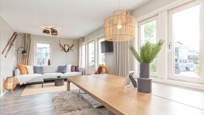 A modern living room with a wooden dining table, a sofa with cushions, a fluffy rug, a potted plant, and a large window with a view of buildings. - Åre Travel- Mitt i Åre 2 (Are)
