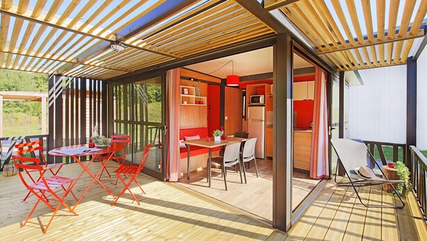 An open-plan living area with a wooden deck, a dining table with red chairs, and a kitchen area with a red appliance.