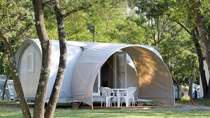 A modern, white, semi-circular tent with a table and chairs in front, surrounded by trees.