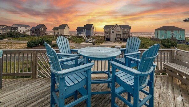 Una terraza de madera con sillas Adirondack azules y una mesa redonda, con vistas a una hilera de casas y al océano durante el atardecer.