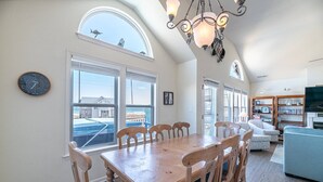 Amplia sala de estar con una gran mesa de comedor de madera, un sofá azul y un televisor. Amplias ventanas ofrecen vistas a la playa y un reloj en la pared.