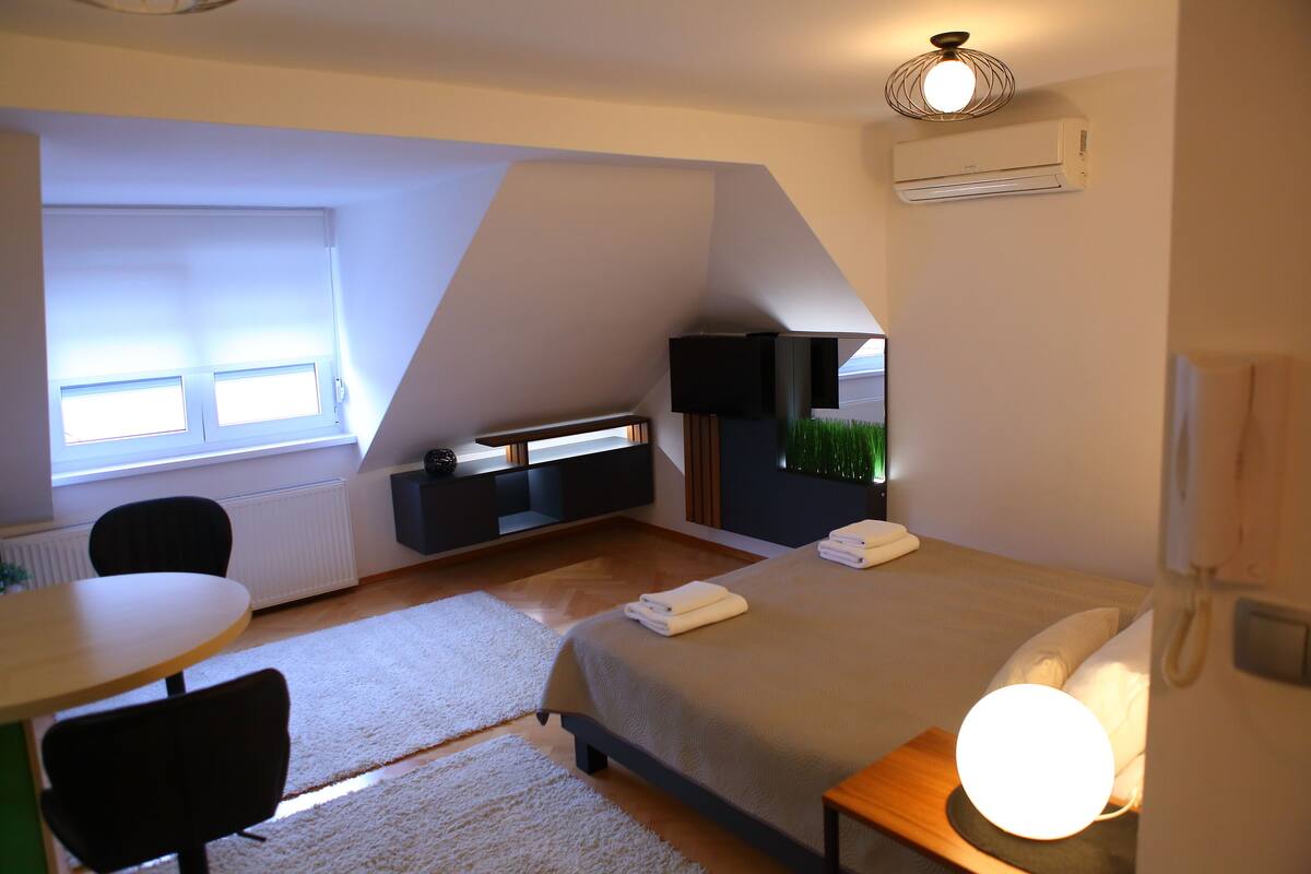 Ein modernes Schlafzimmer mit Bett, Nachttisch und Schreibtisch mit Stuhl. Es gibt ein Fenster mit Jalousien, eine Wand-Klimaanlage und eine Deckenleuchte.