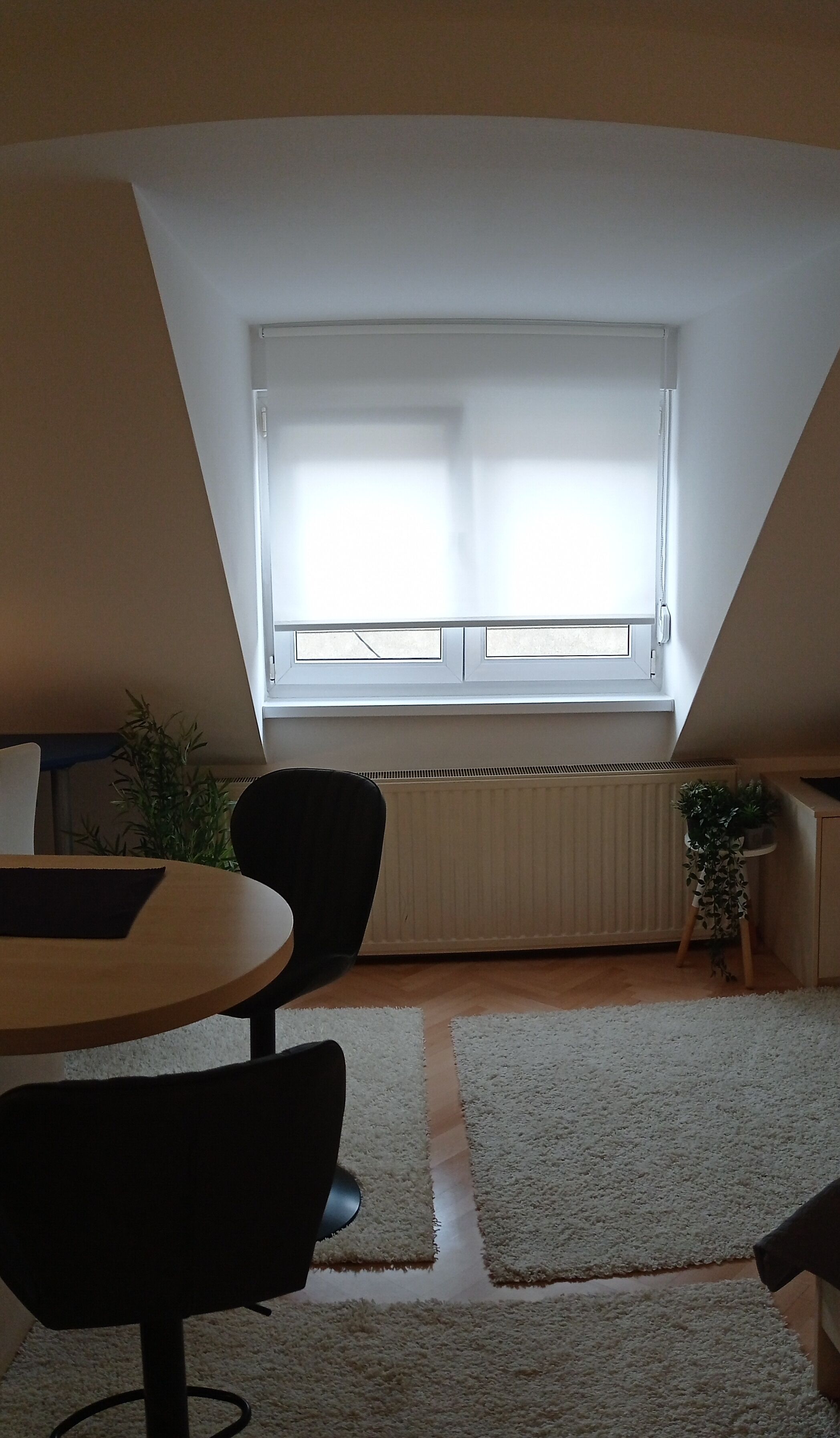 A room with a sloped ceiling, a window with blinds, a radiator, a table with chairs, and a small plant.