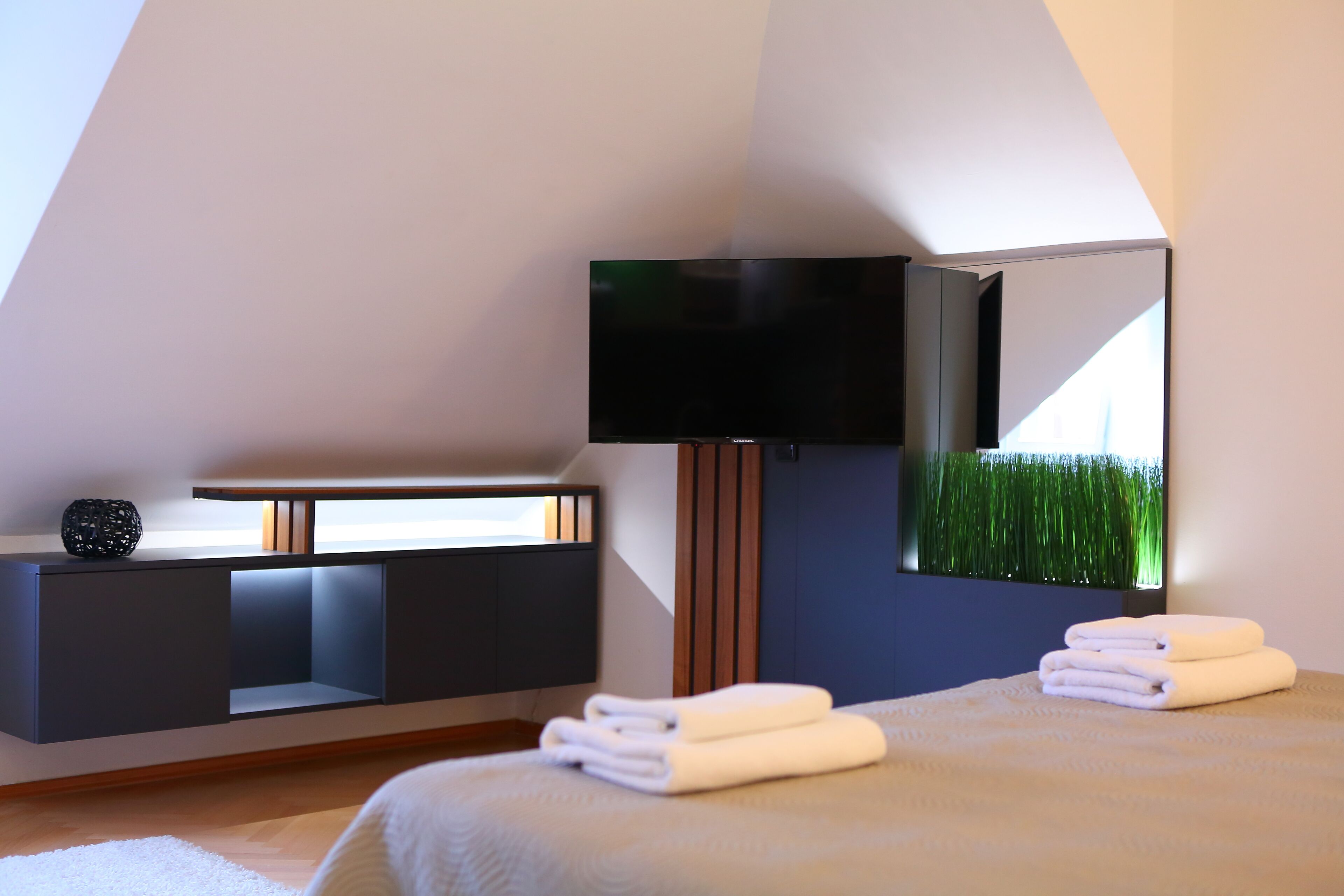 Habitación de hotel moderna con cama, televisor de pantalla plana montado en la pared, estante empotrado para el televisor y un espejo grande que refleja un campo verde.