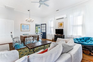 A spacious living room with a dining area, a ceiling fan, a chandelier, a television, and a large window with curtains.