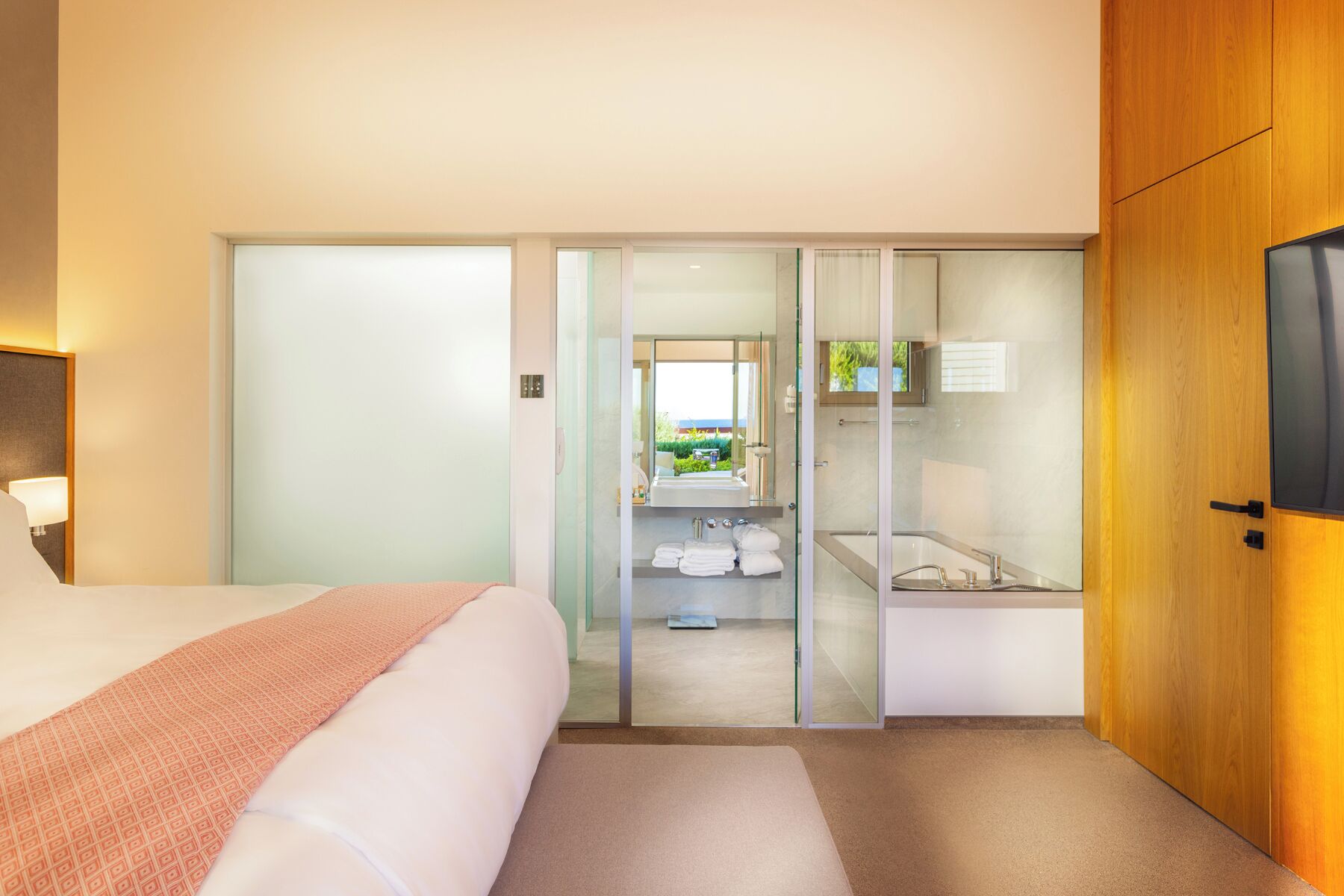 A hotel room with a bed, a glass-enclosed bathroom, a TV, and a wooden door.