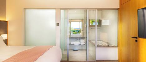 A hotel room with a bed, a glass-enclosed bathroom, a TV, and a wooden door.