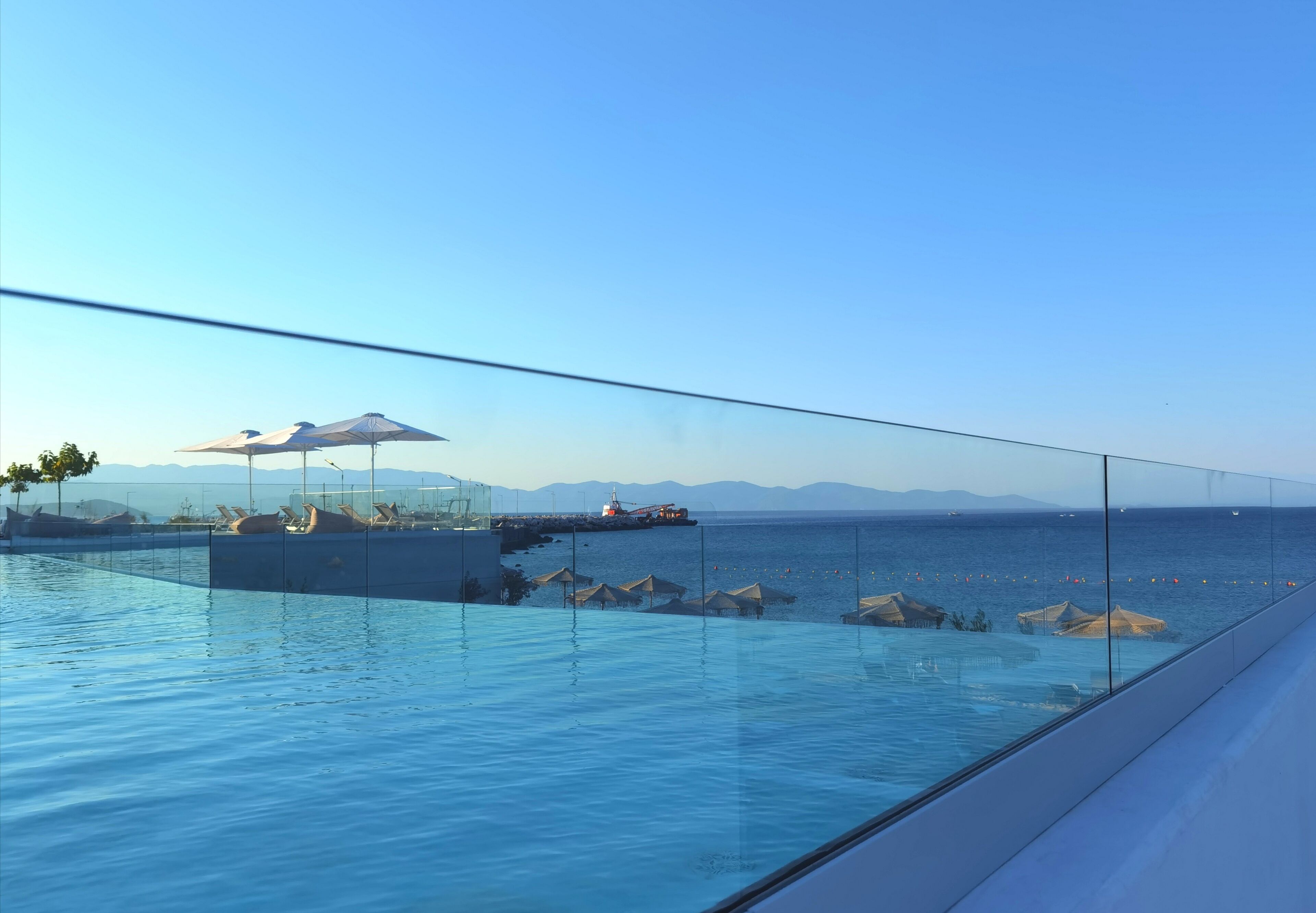 Vista despejada de una piscina infinita con barandilla de vidrio, una zona de descanso con sombrillas y una lejana vista al mar y las montañas.