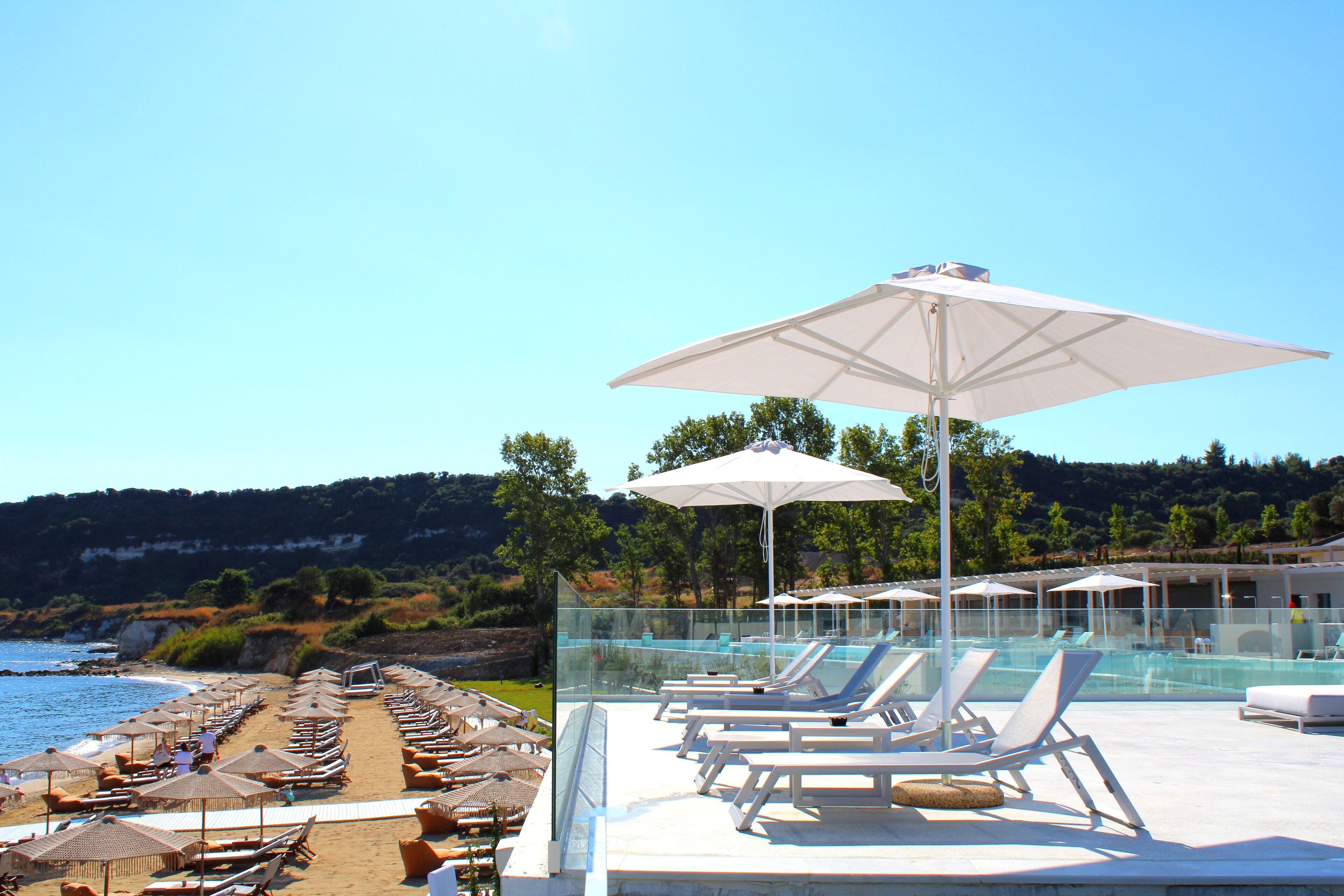 Ein Poolbereich mit Liegestühlen und Sonnenschirmen mit Blick auf einen Strand und einen klaren Himmel.