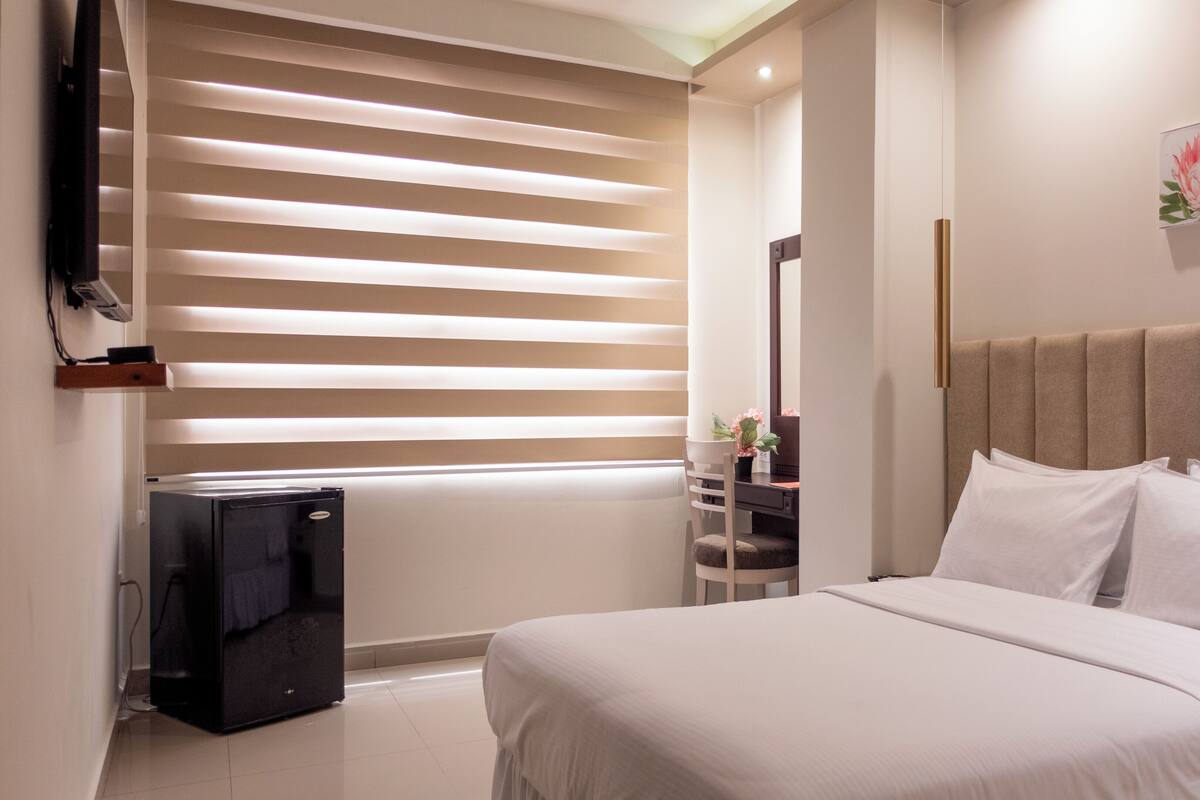 A hotel room with a bed, a desk with a chair, a TV, and a window with blinds.