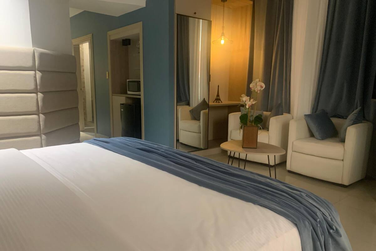 A hotel room with a large bed, a small table with a vase of flowers, two armchairs, a sofa, and a desk with a computer.