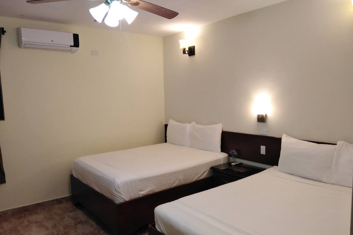 A hotel room with two single beds, a ceiling fan, an air conditioner, and a nightstand with a lamp.