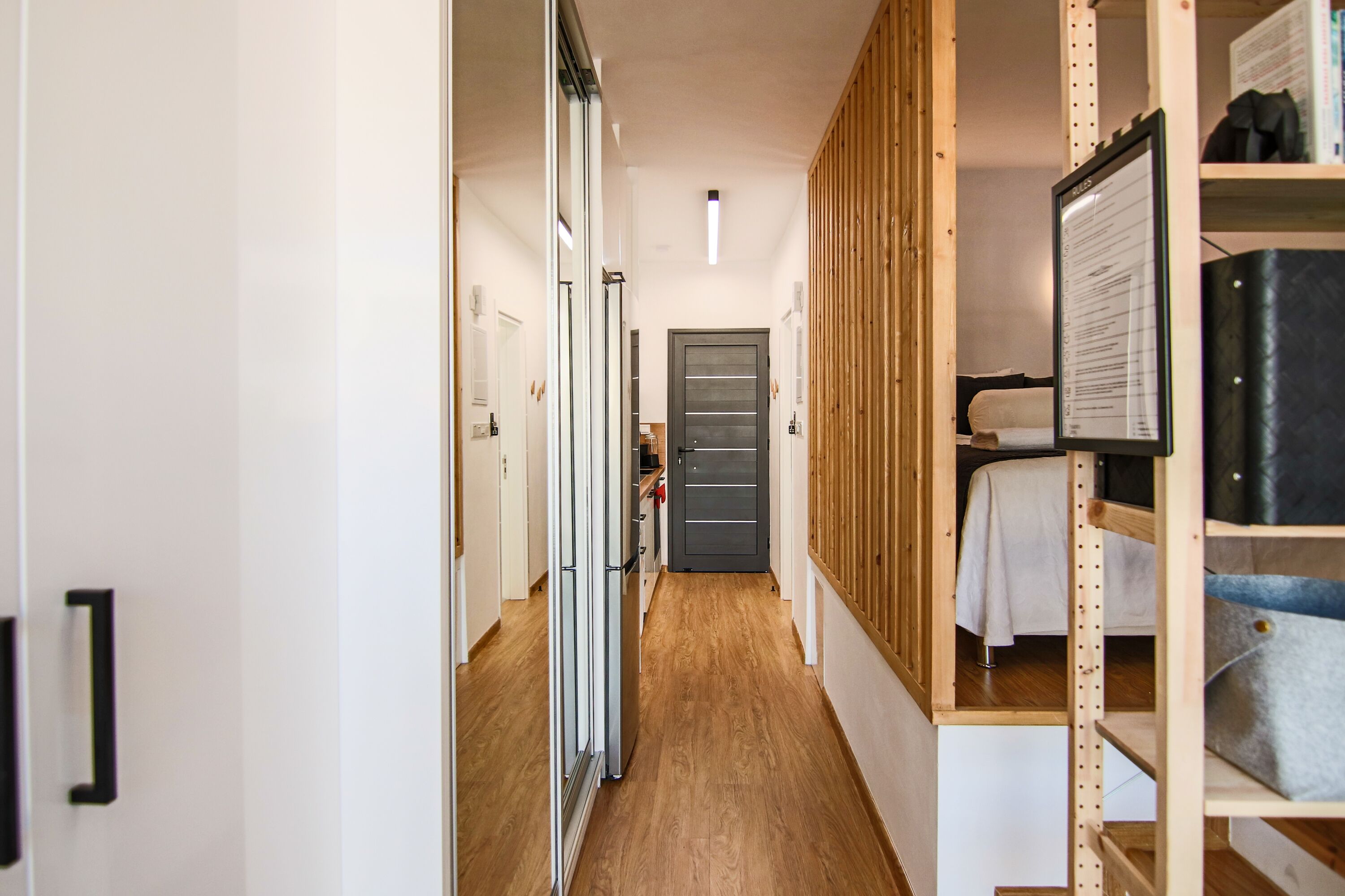 A narrow hallway with a wooden floor, a grey door at the end, and a bed with a white sheet in the background.