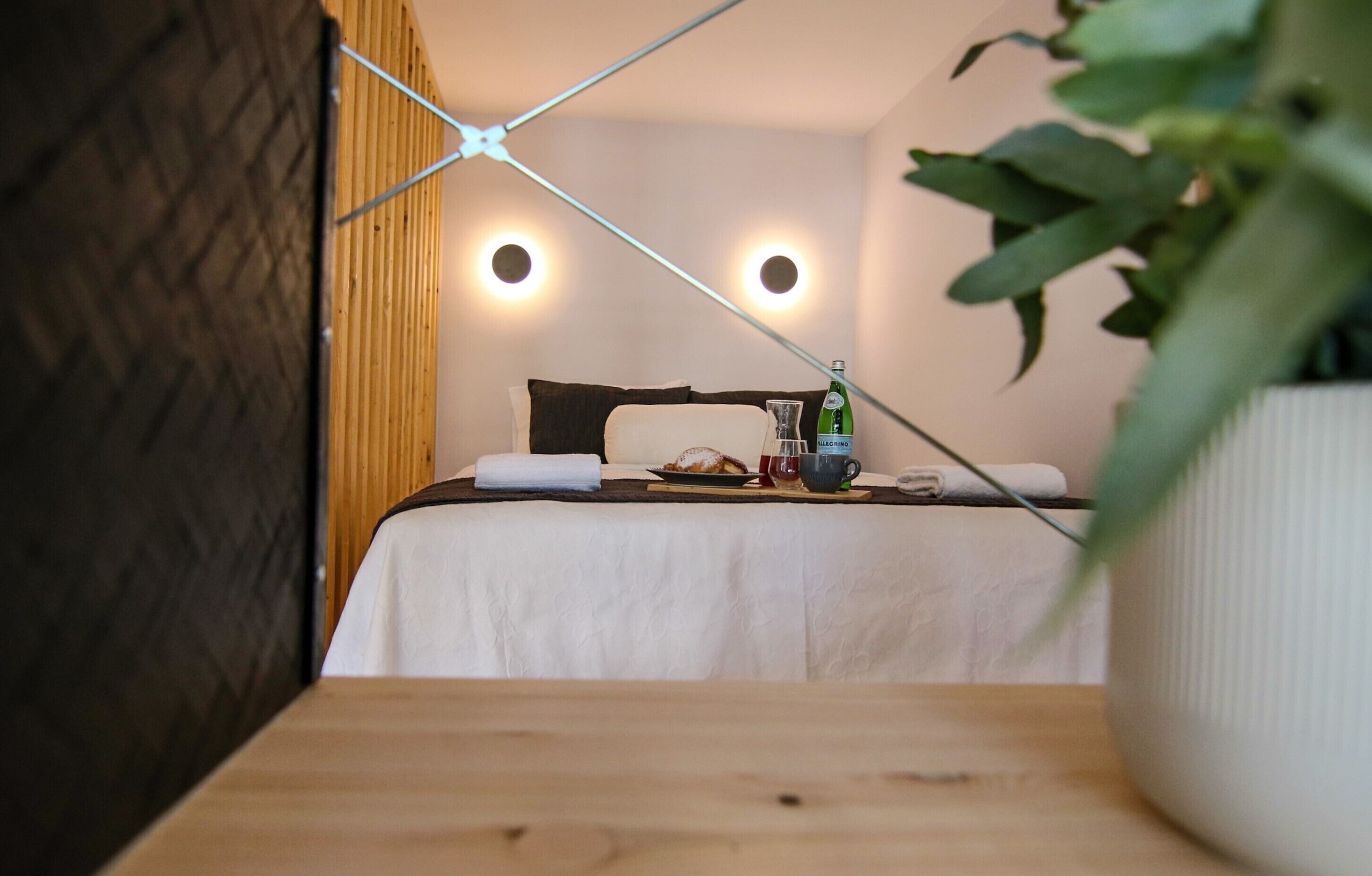 A neatly made bed with a white bedspread, a dark headboard, and a bedside table with a bottle, a glass, and a plate.