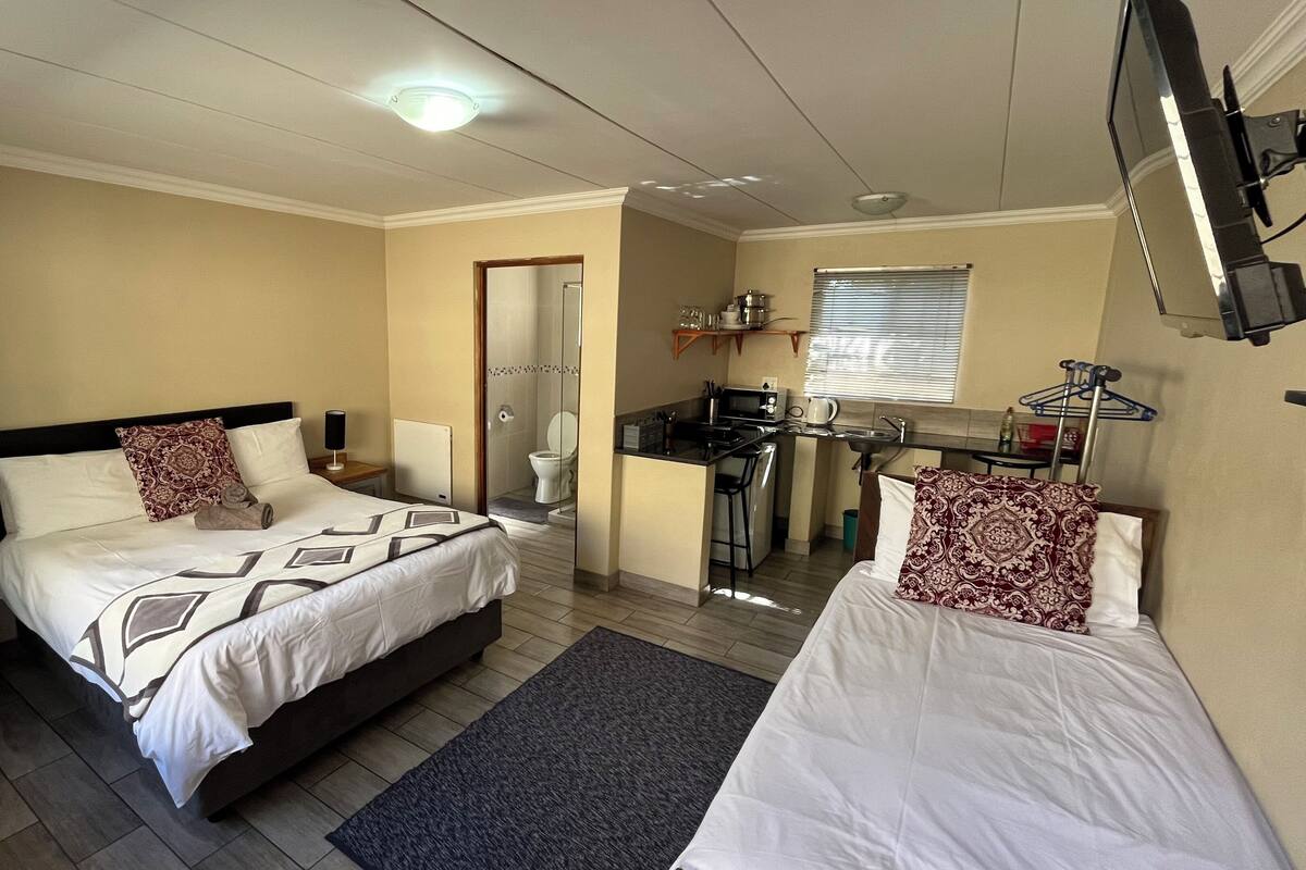 A hotel room with two beds, a small kitchenette, a bathroom with a glass door, and a TV mounted on the wall.