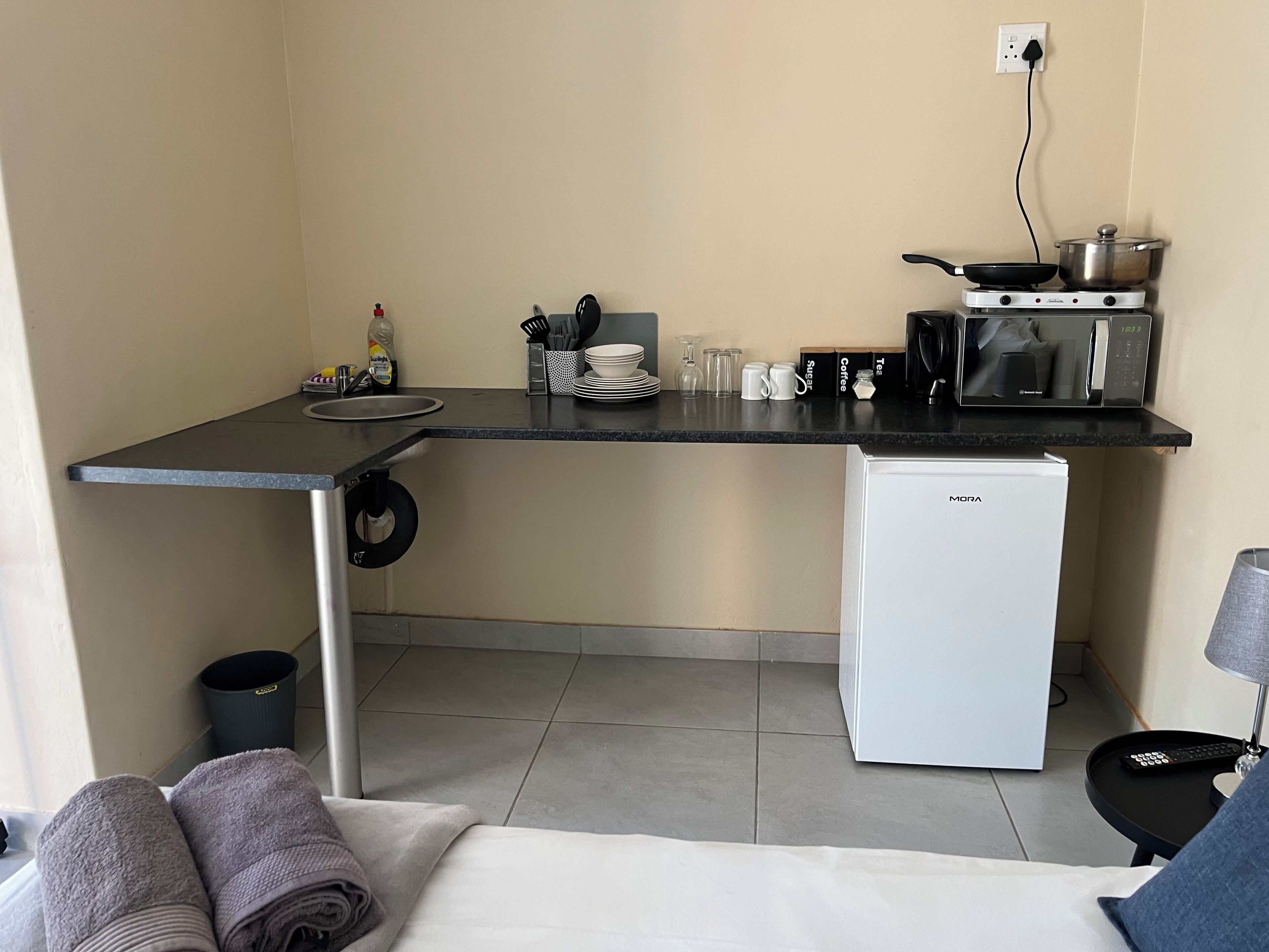 Una kitchenette compacta con estufa, microondas y refrigerador pequeño. Cuenta con un lavamanos y un cubo de basura.