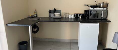 Una kitchenette compacta con estufa, microondas y refrigerador pequeño. Cuenta con un lavamanos y un cubo de basura.