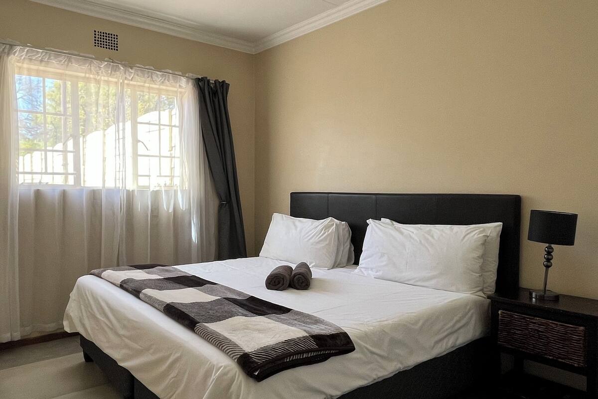A neatly made bed with white linens and a black and white quilt, a bedside lamp, and a window with sheer curtains.