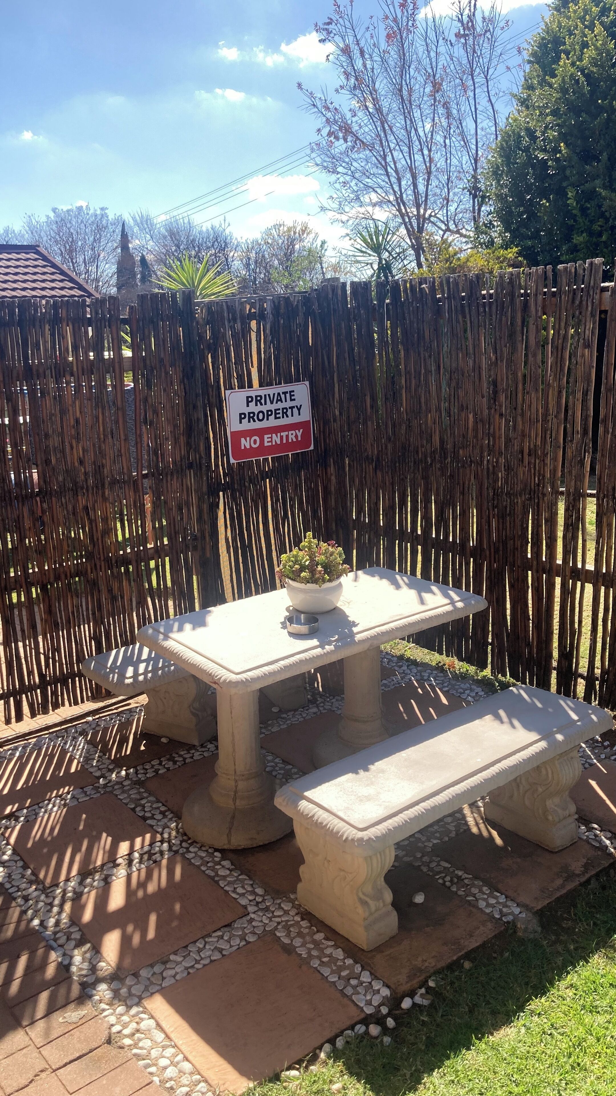 Propriedade privada com placa de 'Proibida a Entrada', um banco de pedra, uma mesa com planta em vaso e um portão decorativo.