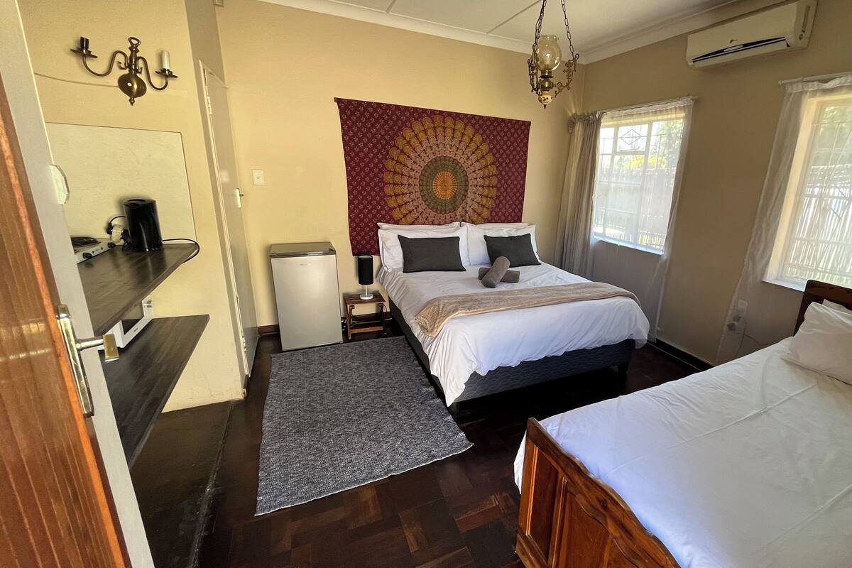 A hotel room with a bed, a desk, a refrigerator, a tapestry, and a window with curtains.