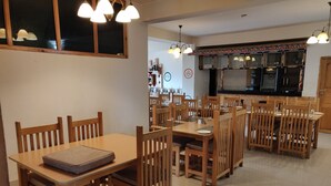 A dining area with wooden tables and chairs, a kitchen in the background, and pendant lighting. - Thori Resort (Thimphu)