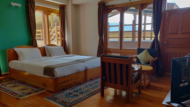 A room with a wooden bed, a chair, a television, and a view of the outdoors through large windows. - Thori Resort (Thimphu)