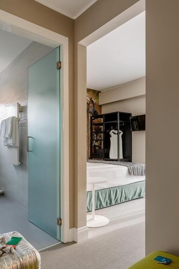 Ett modernt hotellrum med en glasdörr som leder till badrummet, en säng med ett vitt och grönt överkast, ett litet runt bord och en garderob med svart inredning.