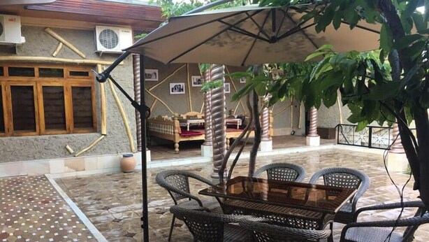 An outdoor patio with a wooden table and wicker chairs, a large umbrella, and a bed in the background. - "S E A S O N S"  (Samarkand)