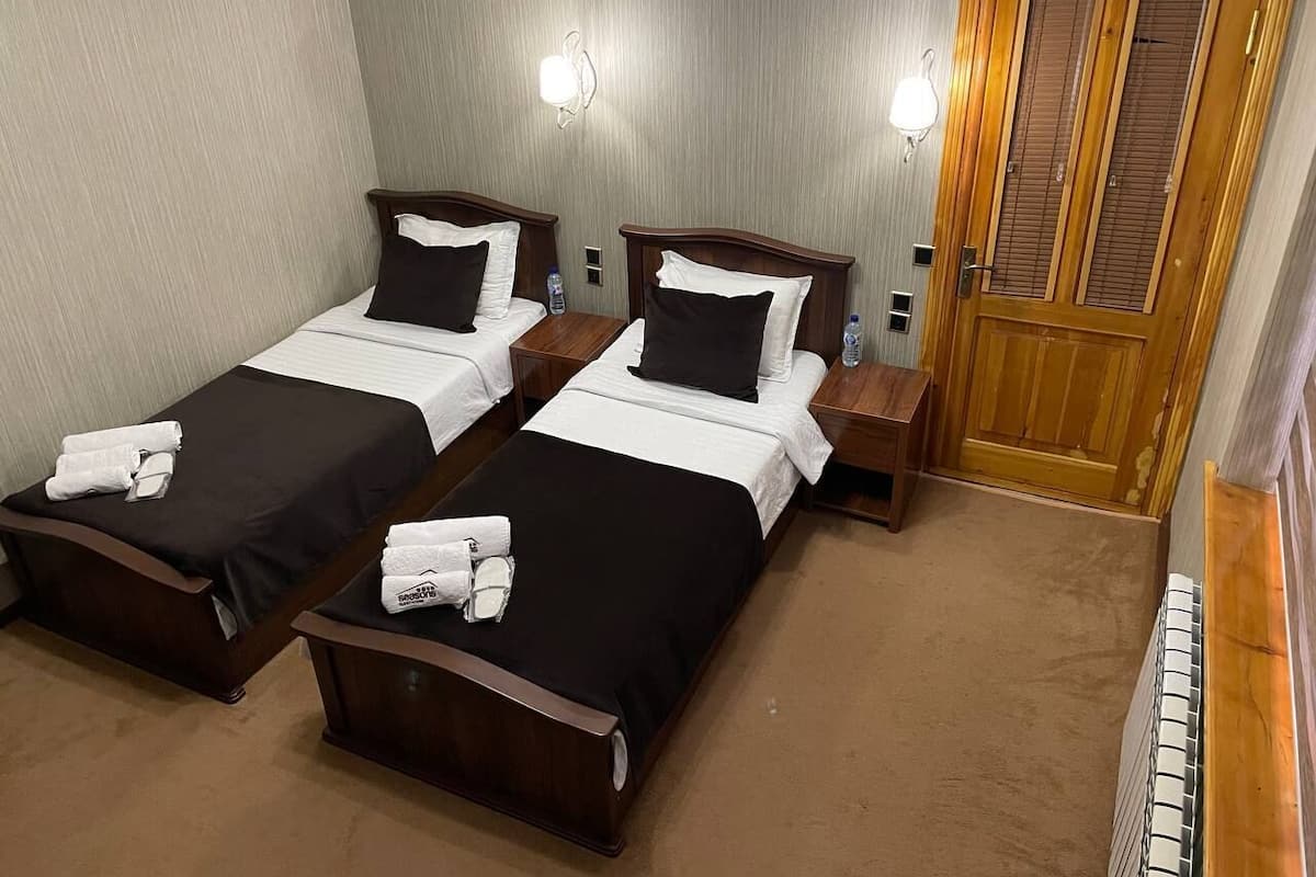 A hotel room with two single beds, each with a wooden headboard, white linens, and a black bedspread. There are two bedside tables with lamps, a wooden door, and a radiator on the right.