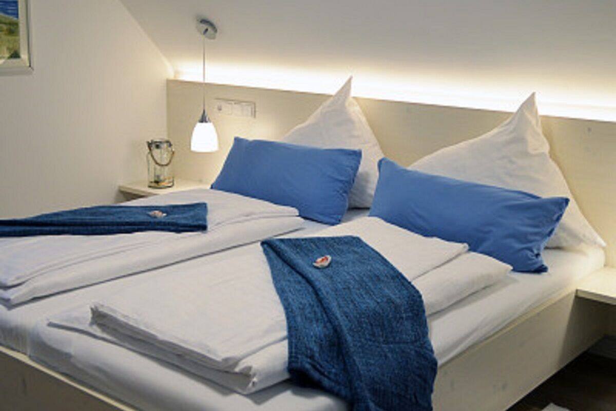 A neatly made bed with white and blue bedding, a bedside lamp, and a small table with a glass container.