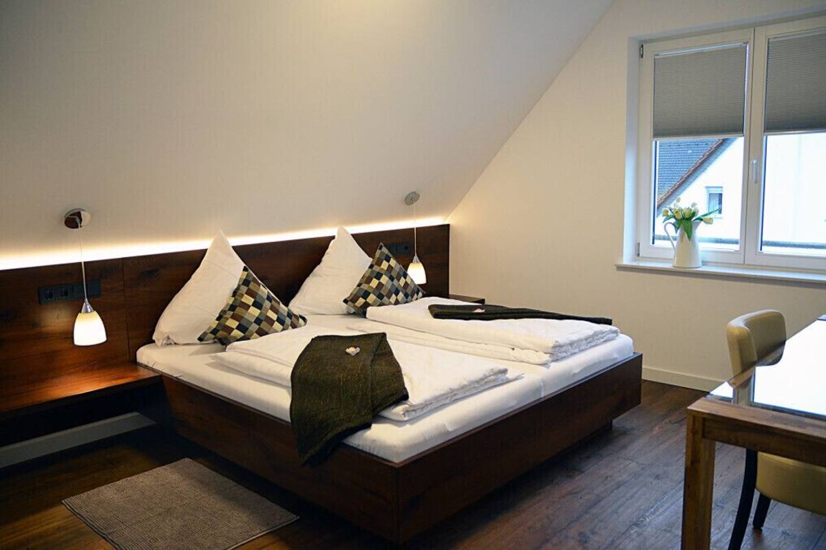A modern bedroom with a wooden headboard, a bed with white linens, a bedside table, a chair, and a window with a view of buildings.