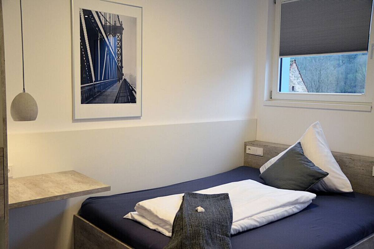 A bedroom with a bed, pillows, a hanging light fixture, a window with a view, and a framed picture on the wall.