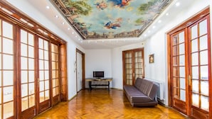 A spacious room with wooden flooring, a sofa, a television, and a ceiling mural.