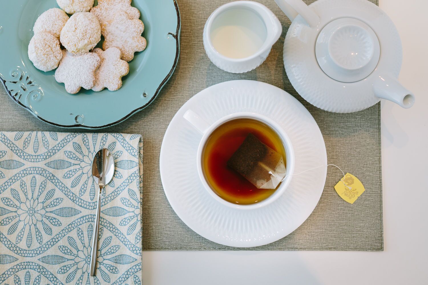 A table setting with a plate of cookies, a cup of tea, a teapot, and a tea bag.