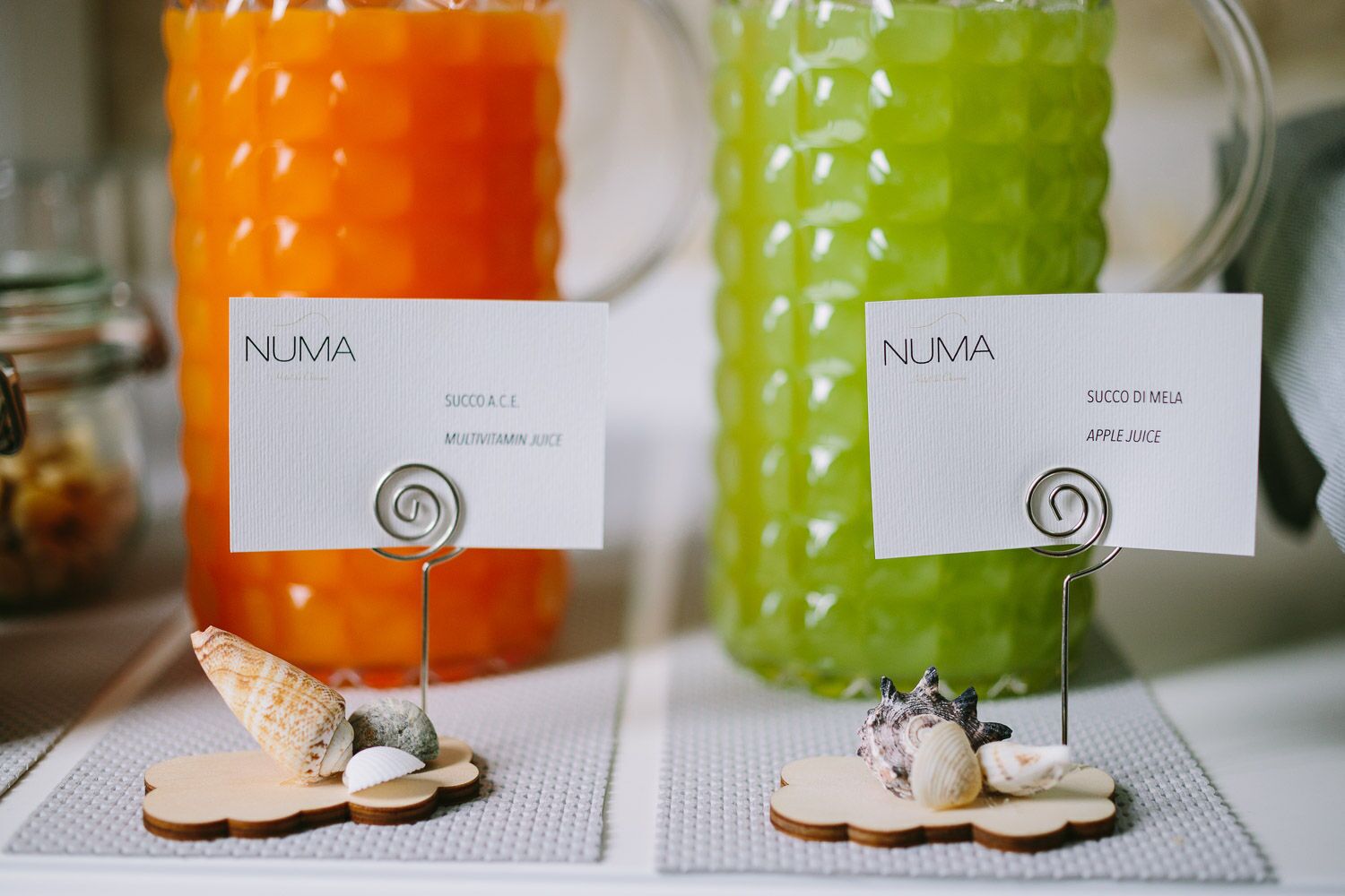 Two labeled juice bottles, one orange and one green, with labels reading "NUMA SUCCO A.C.E. MULTIVITAMIN JUICE" and "NUMA SUCCO DI MELA APPLE JUICE" respectively, placed on a textured surface with seashells and wooden pieces.