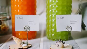Two labeled juice bottles, one orange and one green, with labels reading "NUMA SUCCO A.C.E. MULTIVITAMIN JUICE" and "NUMA SUCCO DI MELA APPLE JUICE" respectively, placed on a textured surface with seashells and wooden pieces.