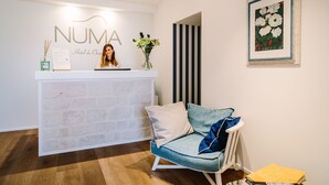 A reception area with a white reception desk labeled "NUMA Hotel de Charité." There is a chair with blue cushions and a framed painting on the wall. - Numa Hotel (Numana)