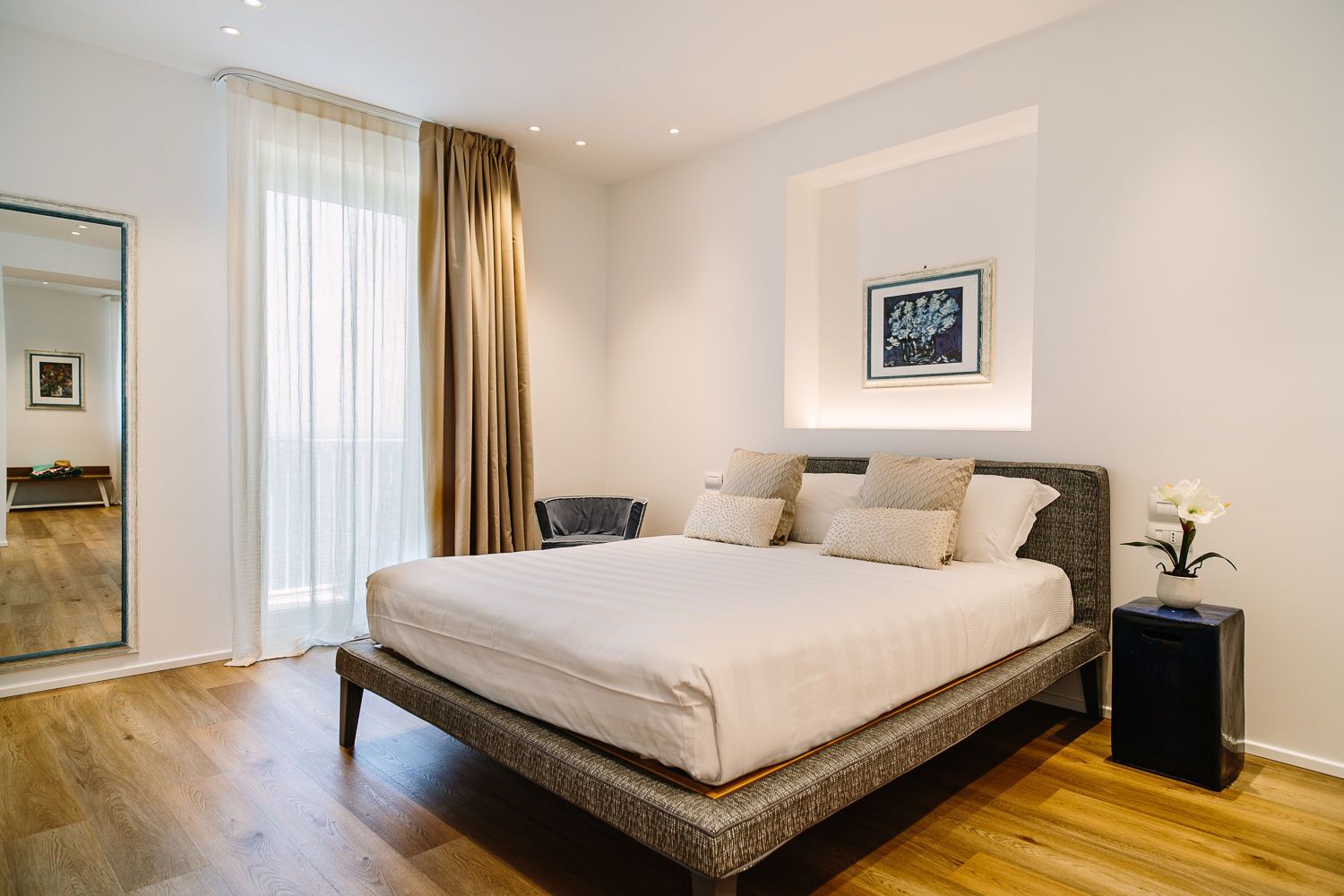 A bedroom with a large bed, a bedside table, a chair, a mirror, and a window with curtains.