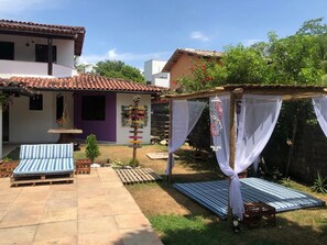 A patio with a blue-striped sofa, a small table, and a hammock. There is a wooden deck and a garden area with a tree and a small bush. - Pé na Areia (Porto Seguro)