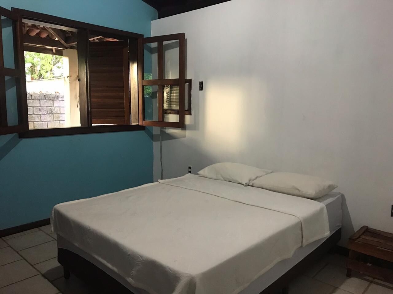 Een enkel bed met een wit matras en kussens, een houten hoofdbord, een raam met houten luiken en een betegelde vloer.