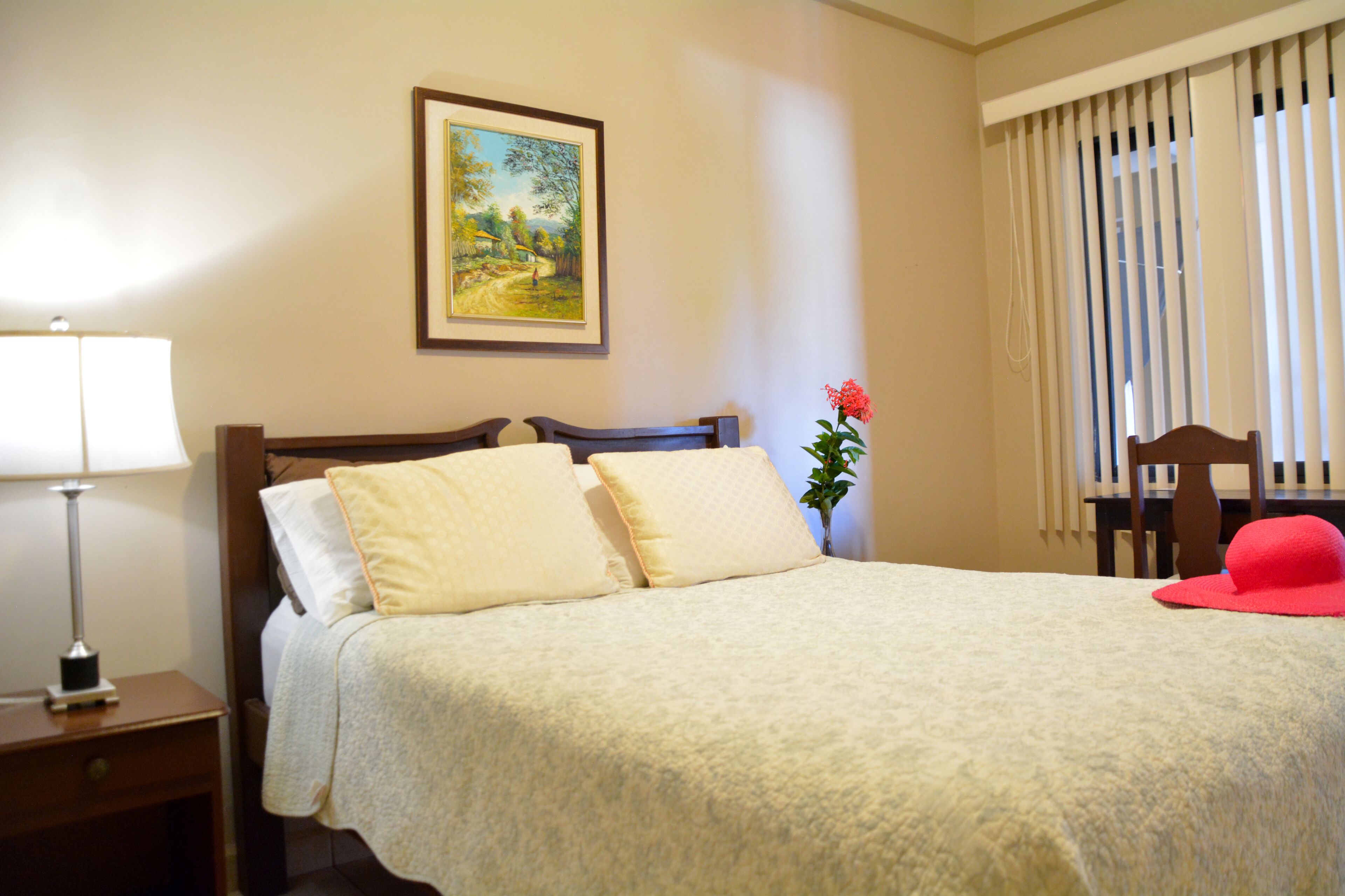 Cama bem arrumada com almofadas, mesa de cabeceira com abajur, quadro na parede, planta em vaso e cadeira com chapéu vermelho.