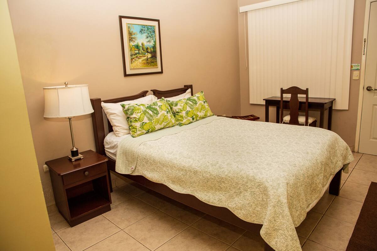 Una cama bien tendida con una colcha estampada con motivos florales y dos almohadas, una mesita de noche con una lámpara, un cuadro de un paisaje en la pared, y una pequeña mesa de madera con una silla.