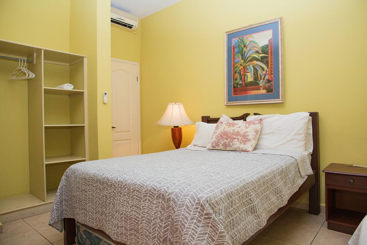 Un dormitorio con cama, mesita de noche, lámpara, armario y un cuadro en la pared.