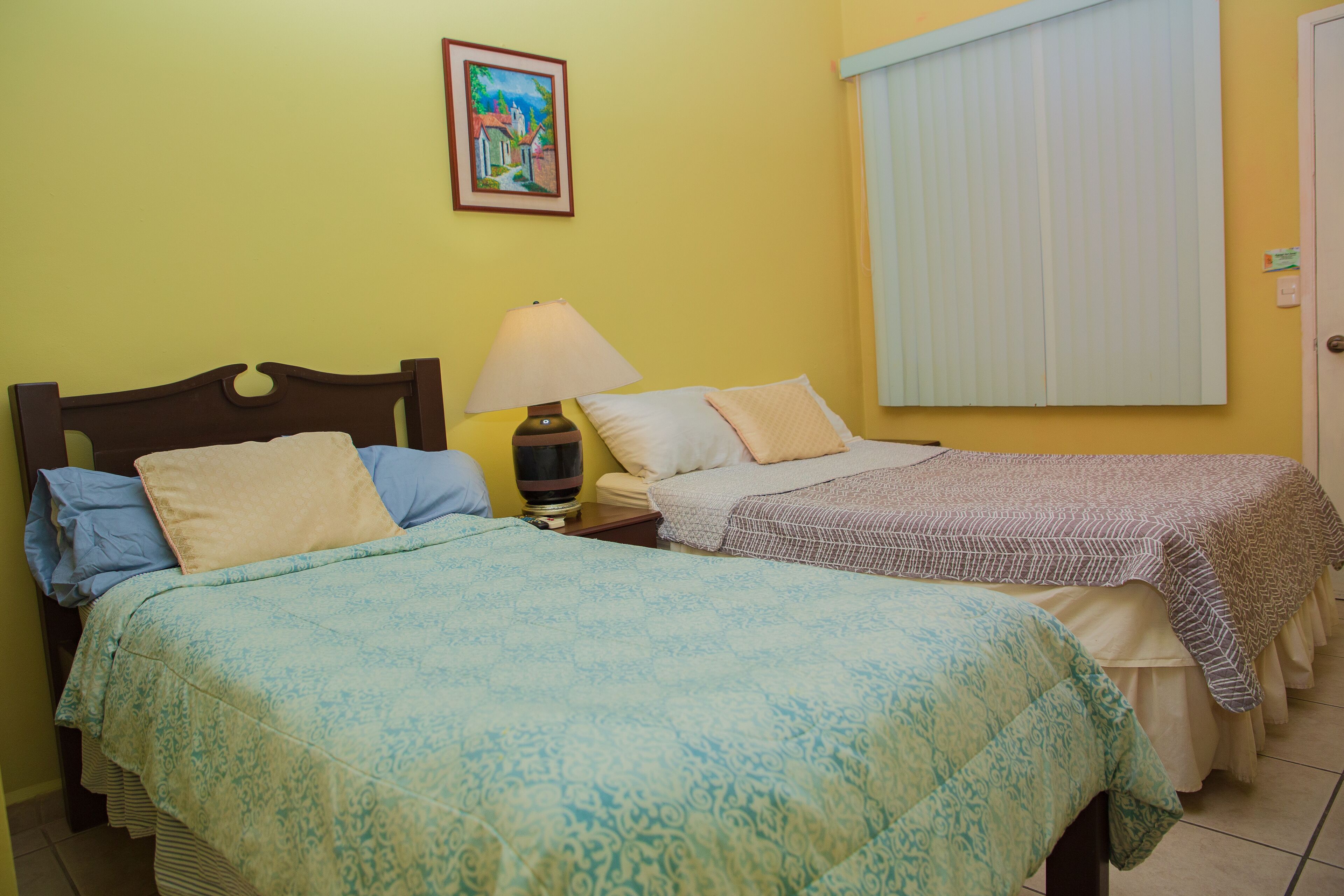 Una habitación con dos camas, cada una con un edredón de estampado diferente, una mesita de noche con lámpara, un cuadro en la pared y una ventana con persianas.