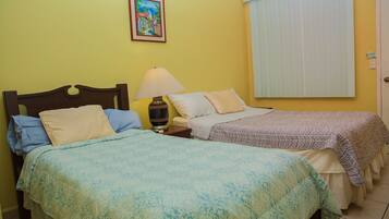 Una habitación con dos camas, cada una con un edredón de estampado diferente, una mesita de noche con lámpara, un cuadro en la pared y una ventana con persianas.