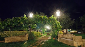 A nighttime garden pathway with stone tiles, illuminated by street lamps, surrounded by greenery and a stone bench. - Aranceto Turismo Rurale (Syracuse)
