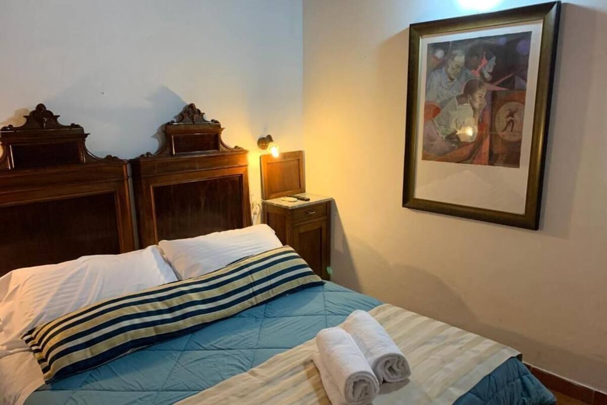 A neatly made bed with striped pillows, a blue quilt, and rolled towels on a wooden headboard. A framed picture hangs on the wall to the right.